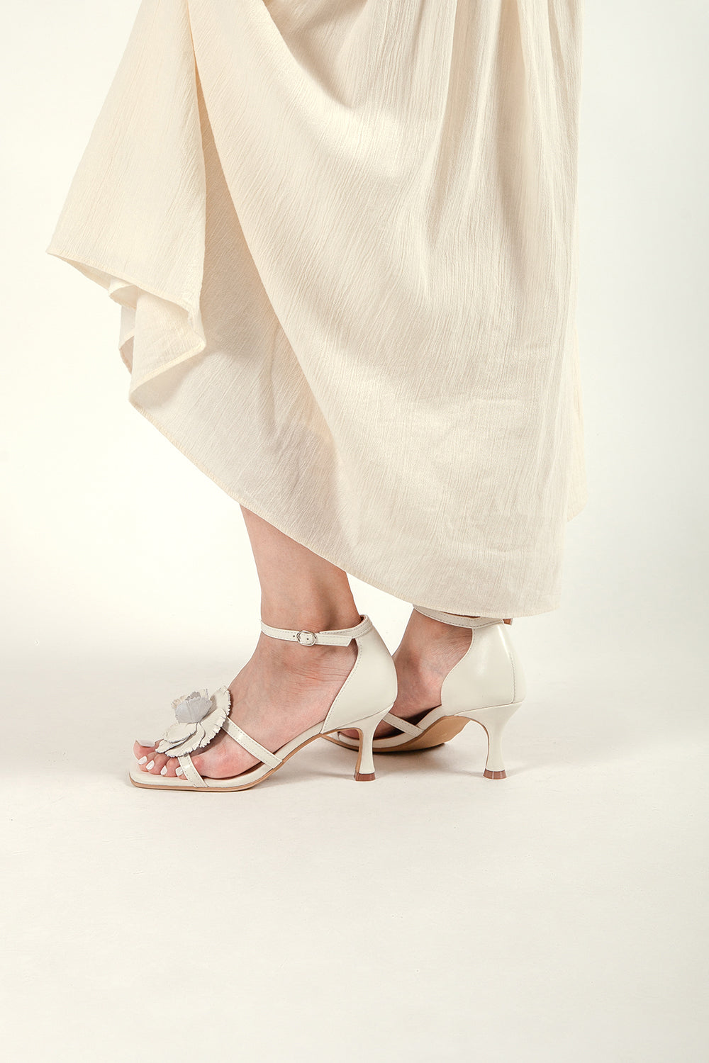 Lifestyle view of a woman wearing ivory floral kitten heel sandals with a flowing cream midi skirt.