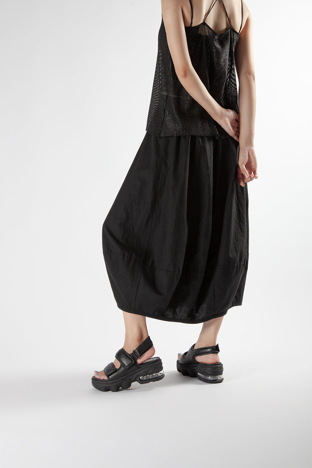 Lifestyle view of a woman wearing black thick-soled air cushion sandals with a black midi skirt and mesh top.